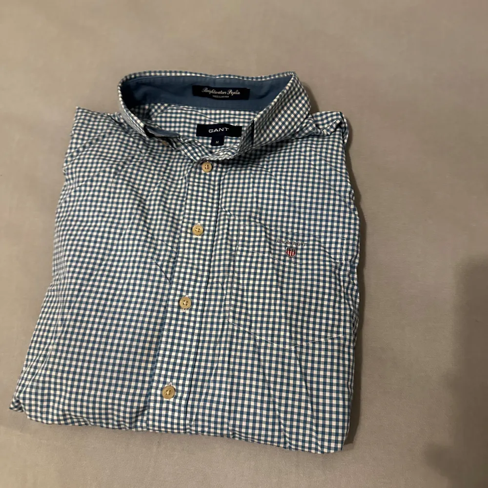 Snygg blårutig skjorta från GANT i storlek M. Skjortan har långa ärmar och klassiska knappar framtill. Perfekt för en stilren look med en liten broderad logga på bröstet. Passar utmärkt till både vardag och mer uppklädda tillfällen.. Paidat.
