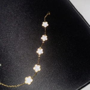 Guldarmband med vita blommor - Elegant guldarmband med fem vita blomformade hängen. Perfekt för att ge en stilren touch till din outfit. Blommorna är jämnt fördelade längs den tunna kedjan. Armbandet har justerbara kedjor som kan anpassa för att passa olika handledsstorlekar. Det finns totalt 2 hål.