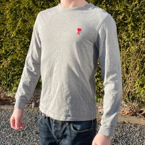 Grå långärmad tröja från Ami - Säljer en stilren grå långärmad tröja från Ami med ett litet rött hjärta broderat på bröstet. Perfekt för en avslappnad look. Tröjan är i mjukt material och har en normal passform. Passar bra till jeans eller chinos.