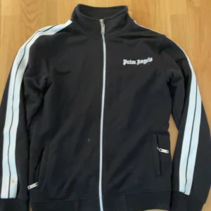 Svart track kofta från Palm Angels - Säljer en svart track jacket från Palm Angels med vita ränder längs ärmarna och dragkedja framtill. Jackan har en hög krage och två fickor med dragkedja. Perfekt för en sportig look eller avslappnad stil.