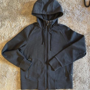 Svart huvtröja från Boss - Snygg svart huvtröja från Boss med dragkedja och justerbar huva. Tröjan har långa ärmar och praktiska fickor framtill. Perfekt för en casual look med stil. Den gröna detaljen på insidan ger en extra touch.