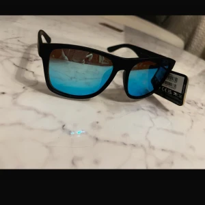 Svarta solglasögon från Foster Grant - Snygga svarta solglasögon från Foster Grant med blå spegelglas. De har en klassisk rektangulär form och polariserade linser för att minska bländning. Perfekta för soliga dagar och ger en cool look.