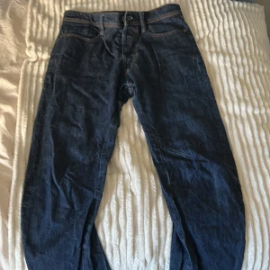 Mörkblå jeans från G-Star RAW - Snygga mörkblå jeans från G-Star RAW med klassisk femficksdesign och orange sömmar. Byxorna har en knappgylf och märkesdetaljer på baksidan. Perfekta för en avslappnad stil.
