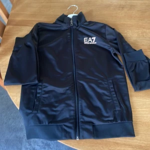 Svart jacka från Emporio Armani EA7 - Säljer en stilren svart jacka från Emporio Armani EA7. Jackan har dragkedja framtill och en hög krage. Perfekt för en sportig look med sin snygga design och bekväma passform.samt byxorna till stället för billigt pris ge gärna bud 