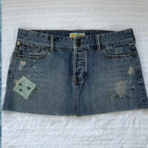 Jeanskjol med pärldetaljer - Trendig jeanskjol med slitna detaljer och pärlbroderier. Kjolen har en kort längd och rå kant nedtill. Y2K, går inte att få tag på längre!!! Helt ny. Storlek 9 (27/28?). Midjemått 82 cm, framsidans längd 28 cm, baksidans längd 31 cm. Priset går inte att diskutera, använd gärna KÖP NU.