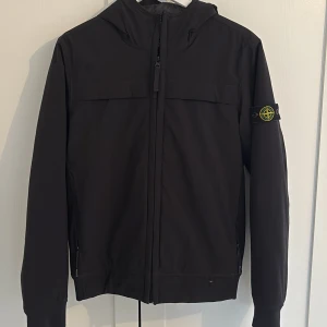 Svart jacka från Stone Island Junior - Säljer en svart jacka från Stone Island Junior med dragkedja och broderad logga på ärmen. Jackan har långa ärmar, och är fodrad. Perfekt för den som gillar en klassisk look. Litet hål som syns på bild 2. I övrigt mycket fint skick. Självklart äkta, inköpt på nk kids i Stockholm.