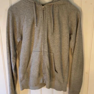Grå hoodie i ekologisk bomull - Säljer en grå hoodie i ekologisk bomull med dragkedja och huva. Tröjan har långa ärmar och ribbade muddar. Perfekt för en avslappnad stil. Tröjan är trasig vid slutet av ärmen, något jag aldrig stört mig på.