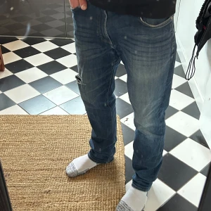 Blå jeans med slitningar - Snygga blå jeans med en cool slitning på ena benet. De har en klassisk femficksdesign och en knappgylf. Perfekta för en avslappnad stil. W32 L32. 