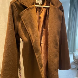 Brun kappa från NA-KD - Snygg brun kappa från NA-KD med klassisk design och knytbälte i midjan. Perfekt för höst och vår, Passar både till vardags och mer uppklädda tillfällen.