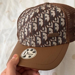 Brun keps från Dior - Snygg brun keps från Dior med ett unikt mönster i beige och brunt. Kepsen har ett nätmaterial på sidorna och en brun skärm. Perfekt för en stilren look.