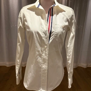 Vit skjorta från GANT - Snygg vit skjorta från GANT med klassisk krage och broderad logga. Skjortan har långa ärmar och en bröstficka med dekorativa ränder i rött och blått. Perfekt för en stilren look.