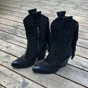 Svarta cowboy boots med nitar och fransar - Riktigt snygga svarta cowboy boots med spetsig tå, klack och massor av svarta nitar. Skorna har fransar där bak och dragkedja för enkel påtagning. Både i skinn och mocka. Precis behandlade och impregnerade, däremot lite kantstötta vid tån. Endast använda vid tre tillfällen. Låda saknas. 