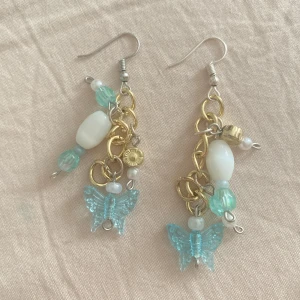 Örhängen med blå fjärilar och pärlor - Unika örhängen med blå fjärilshängen i plast, guldfärgade kedjor och detaljer, vita pärlor och turkosa glaspärlor. Mixen av material och färger ger en lekfull och trendig vibe. Perfekta för dig som gillar statement-accessoarer och vill sticka ut.