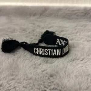 Svart armband från Christian Dior - Snyggt svart armband från Christian Dior med vit broderad logga. Armbandet har tofsar i ändarna och är gjort i ett mjukt textilmaterial. Perfekt accessoar för att lyfta din outfit med en lyxig touch.