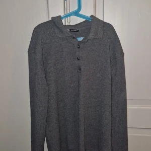 Grå ribbad polotröja H&M - Mysig grå ribbad polotröja från H&M med knappar framtill och klassisk krage. Långärmad modell i mjukt stickat material, perfekt för lager-på-lager och chill dagar.