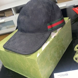 Svart Gucci keps med logga och ränder - Säljer en svart keps från Gucci med diskret GG-monogram över hela kepsen och klassiska gröna och röda ränder på sidan. Justerbar rem baktill och tillverkad i bomull. Perfekt för dig som vill ha en lyxig och stilren accessoar. Såklart äkta och du får med alla tillbehör direkt! Pris kan diskuteras vid snabb affär