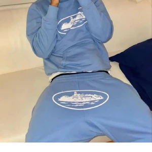 Ljusblå hoodie med skeppstryck - Säljer en ljusblå hoodie med huva och magficka. På bröstet finns ett stort vitt tryck av ett skepp i oval ram. Hoodien är i mjukt material och har en avslappnad passform, perfekt för chill dagar.