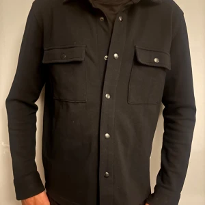 Svart overshirt med tryckknappar - Stilren svart overshirt med raka linjer och två stora bröstfickor med lock och tryckknappar. Skjortan har klassisk krage och långa ärmar. Perfekt att bära öppen eller stängd för en clean look.