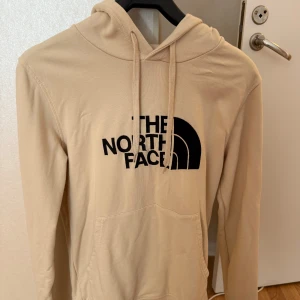 Beige hoodie från The North Face - Säljer en beige hoodie från The North Face med svart logga framtill. Tröjan har huva med snörning och en stor magficka. Långärmad och tillverkad i mjuk bomull, perfekt för chill dagar eller när du vill ha en clean look.