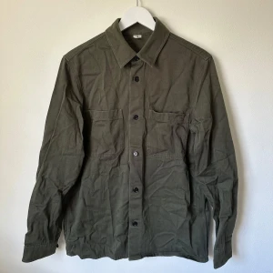 Olivgrön overshirt från Arket - Olivgrön overshirt från Arket i bomull med klassisk krage, två stora bröstfickor och svarta knappar framtill. Långärmad modell med knappslå vid ärmslut. Perfekt för lager-på-lager och chill stil. C383