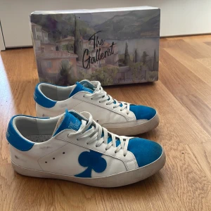 The Gallerist vita och blå sneakers - Snygga vita sneakers från The Gallerist med blå detaljer och klubbmärke på sidan. Skorna har blå mockadetaljer på tå och plös, samt snörning framtill. Tillverkade i skinn och mocka för en cool och unik look. Skorna är extremt billiga för sitt skick, så först till kvarn. Skriv vid minsta fundering!