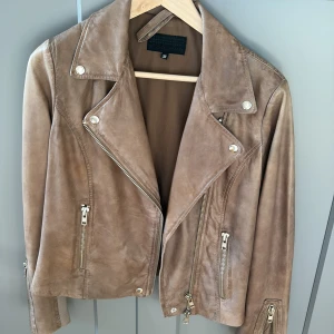 Beige skinnjacka från Frontrow, stl 38 - Säljer en snygg beige skinnjacka från Frontrow i storlek 38. Jackan har klassisk bikerstil med silvriga dragkedjor, bred krage och coola detaljer. Tillverkad i 100% skinn med mjukt foder. Perfekt för dig som vill ha en tidlös och trendig look.