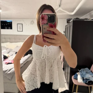 Vit spetsig linnetopp med knyt - Supersöt vit linnetopp med spetsdetaljer och volangig nederdel. Toppen har smala axelband och knyt framtill, vilket ger en romantisk och luftig känsla. Perfekt för varma dagar och somriga outfits.