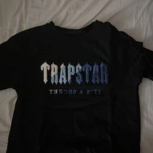Svart Trapstar t-shirt med blått tryck - Svart t-shirt från Trapstar med stort tryck på bröstet i blå och vit gradient. Klassisk passform med rund hals och korta ärmar. Perfekt för dig som gillar streetwear och vill sticka ut med en fet logga.