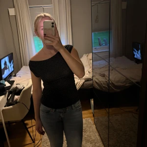 Svart offshoulder prickig topp - Säljer en svart offshoulder topp med små vita prickar. Toppen har en tight passform och ärmlös design med asymmetrisk axel. Perfekt för dig som gillar en enkel men ändå lite edgy stil. Materialet känns mjukt och följsamt mot kroppen.