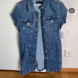 Kort jeansklänning med knappar - Snygg kort jeansklänning från Only med korta ärmar, klassisk skjortkrage och knappar hela vägen framtill. Två bröstfickor med lock och knappdetaljer ger en cool look. Perfekt för dig som gillar denim och vill ha en avslappnad stil.