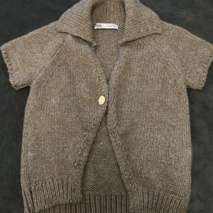 Beige stickad kofta från Zara - Kortärmad stickad kofta från Zara i beige nyans. Koftan har en bred krage, en guldfärgad knapp framtill och ribbade muddar. Perfekt att slänga över en t-shirt för en chill och snygg look.