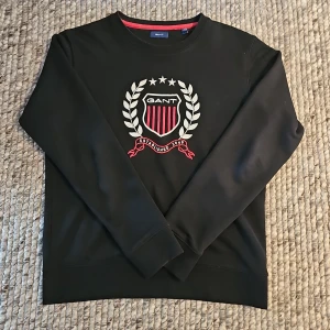 Svart sweatshirt med broderad GANT-logga - Svart sweatshirt från GANT med broderad sköld och vita detaljer framtill. Tröjan har rund hals, långa ärmar och ribbade muddar. Snygg röd och vit brodyr ger en sportig och klassisk vibe. Perfekt för dig som gillar stilrena och preppy plagg.