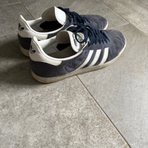 Adidas Gazelle sneakers mörkblå mocka - Säljer ett par klassiska Adidas Gazelle sneakers i mörkblå mocka med vita detaljer och snörning. Skorna har de ikoniska tre ränderna på sidan, rund tå och platt sula. Adidas-logga på hälen och tungan. Köpta för 1000kr på Zalando. Och är i okej, bra skick då de är lite smutsiga!