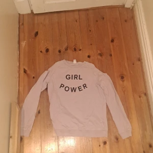 Ljuslila sweatshirt med GIRL POWER-tryck - Ljuslila sweatshirt med glittrig marinblå text 'GIRL POWER' på bröstet. Tröjan har rund halsringning, långa ärmar, ribbade muddar vid ärmslut, hål på axlarna och nederkant. Perfekt för dig som vill ha en enkel men statement tröja i garderoben.