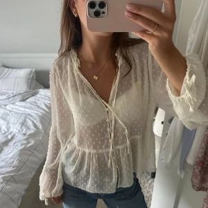 Vit transparent blus med knyt och prickar - Superfin vit blus i transparent material med små prickar, knytning vid halsen och vida ärmar med rosett-detaljer. Blusen har knappar framtill och volang nedtill för en romantisk vibe. Perfekt att styla med jeans eller kjol.