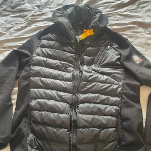 Parajumper cardigan - Svart cardigan jacka från Parajumpers med ikonisk gul rem vid kragen och patch på ärmen. Jackan har quiltad front, huva och dragkedja hela vägen. Ärmarna är i mjukare material för extra komfort. Perfekt för dig som vill ha en snygg och funktionell jacka. Aldrig använd är i helt nytt skick. Säljer då den inte kom till användning. Priset kan diskuteras