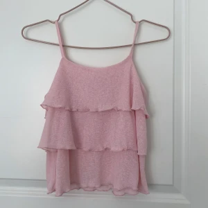 Rosa volang-linnetopp från Kappahl - Supersöt ljusrosa linnetopp från Kappahl med tunna axelband och flera lager volanger. Toppen är ärmlös och har en flowy passform som känns både luftig och trendig. Perfekt för varma dagar när du vill ha en söt och enkel look.