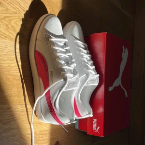 Vita och rosa sneakers från Puma - Snygga vita sneakers från Puma med klassisk design och rosa sidodetalj. Skorna har snörning, rund tå och platt sula. Tillverkade i syntetmaterial med bekväm innersula. Perfekta för dig som gillar stilrena och sportiga vibes.