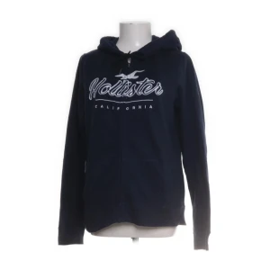 Marinblå hoodie från Hollister zip up - Snygg marinblå hoodie från Hollister med vit logga och text på bröstet. Tröjan har huva med snörning, dragkedja framtill och två fickor. Perfekt för chill dagar eller när du vill ha en avslappnad stil.