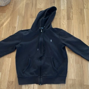 Svart hoodie från Polo Ralph Lauren - Svart hoodie från Polo Ralph Lauren med klassisk logga på bröstet. Tröjan har huva med snörning, dragkedja framtill och två stora fickor. Materialet är mjuk bomull och passformen är normal. Storlek S passar xs