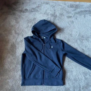  Polo Ralph Lauren - Snygg marinblå hoodie från Polo Ralph Lauren med dragkedja och huva med snörning. Inga skador eller liknade. Storlek M men sitter som S.Vid fundering hör gärna av dig.