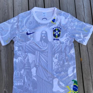 Vit Brasil Nike fotbollströja S - Säljer en vit Brasilien fotbollströja från Nike med detaljerat tryck av Kristusstatyn och brasilianska motiv. Tröjan har korta ärmar, CBF-emblem på bröstet och inslag av blått, gult och grönt. Materialet är lätt polyester som andas, perfekt för match eller träning.