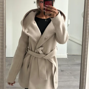 Beige kappa med huva och bälte - Snygg beige kappa med stor huva och knytskärp i midjan. Jackan har knappar framtill och långa ärmar, vilket ger en chill och stilren look. Perfekt för dig som gillar neutrala färger och vill ha en jacka som sticker ut med sin breda krage. Jag har storlek 32 och den passar mig väldigt bra.