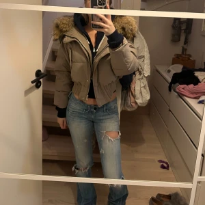 Beige croppad pufferjacka med äkta päls - Säljer en beige croppad pufferjacka med stor äkta päls. Jackan har dragkedja och knappar framtill, bred resår i midjan och långa ärmar. Perfekt för kalla dagar när du vill ha en snygg och trendig look. 