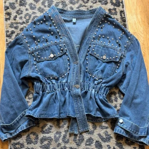 Snygg denimjacka med nitar & svarta strass stenar  - Riktigt snygg och annorlunda denim jacka med massor av silverfärgade nitar och svarta strasstenar Det är en lite kortare modell med insvängd midja med töjbar resår Jackan är v-ringad Den har oxå 2st bröstfickor med knapp och långa ärmar med klaff & knapp Det är en mellanblå färg på den och den är tillverkade denim/jeanstyg Mått: L55/Bredd under ärm 50x2-Midjan är töjbar Passar storlek 36-40 (jag är en storlek 38) Den är som ny i jättefint skick!