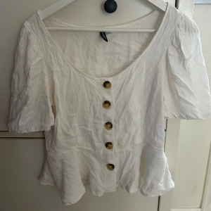 Vit blus med knappar från H&M - Supersöt vit blus med kort puffärm och snygga bruna knappar framtill. Blusen har en lätt v-ringning och volang nertill som ger en romantisk vibe. Perfekt till jeans eller kjol för en fräsch look.