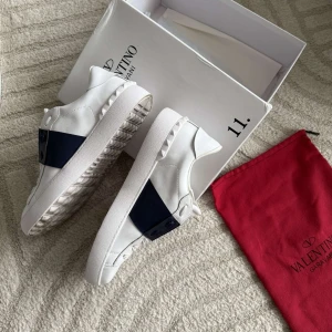 Valentino Garavani opens vita/blåa sneakers - Snygga vita Valentino Garavani sneakers med bred marinblå läderdetalj över snörningen. Skorna har platt sula med coola nitar längs kanten och rund tå. Lyxig design med logga på tungan och klassisk siluett. Perfekt för dig som vill sticka ut med stil. Använd nån gång.