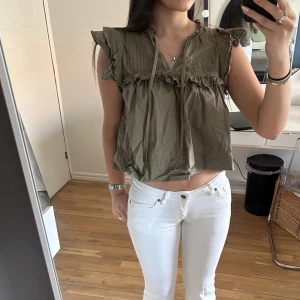 Olivgrön volangblus med knytband - Supersöt olivgrön blus med volangdetaljer och knytband vid halsen. Blusen är ärmlös med rynkade axlar och har en luftig, croppad passform. Perfekt för varma dagar och ger en avslappnad men trendig vibe. Från gina