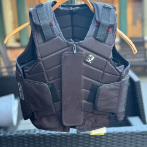 Svart säkerhetsväst L3 från HORKA - Svart ridväst från Horze med quiltad design och justerbara axelband. Västen har dragkedja framtill, breda kardborreband i midjan och reflekterande detaljer längs sidorna. Strl CHILD XL se bild för storleksguide.  