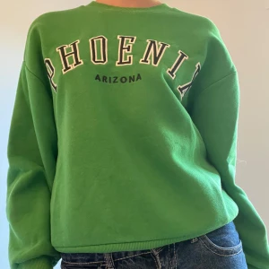 Grön tröja med PHOENIX-tryck - Cool grön sweatshirt / tröja  från Gina Tricot med stort PHOENIX ARIZONA-tryck i vitt och svart på bröstet. Tröjan har rund hals, ribbade muddar och lång ärm. Perfekt för dig som gillar college-vibe och vill sticka ut med färg.